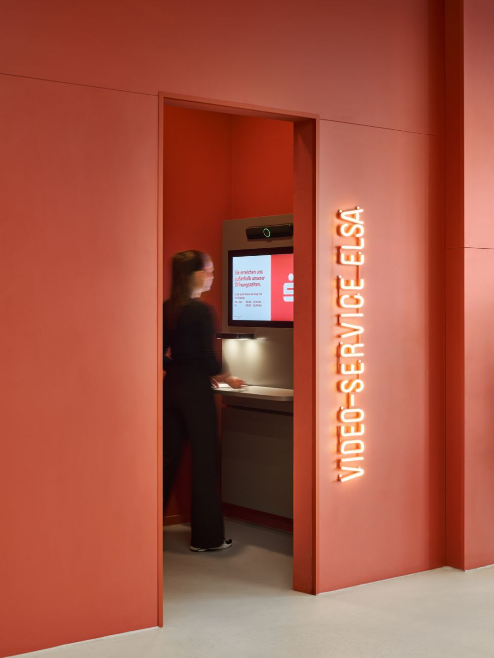 Eine Person nutzt einen Videoservice-Terminal in einer modernen Bankfiliale mit orangefarbenem Design. Beispiel für innovative Filialkonzepte.